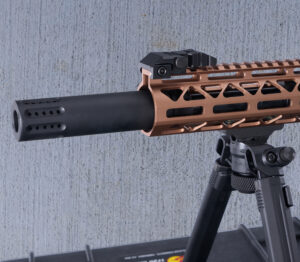 Close-up of modern firearm with suppressor and bronze handguard on bipod.