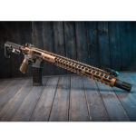Bronze tactical rifle on rustic wooden floor with adjustable stock and muzzle brake.