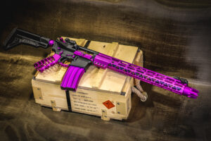 Purple-customized tactical rifle displayed on a wooden crate in a rustic setting.